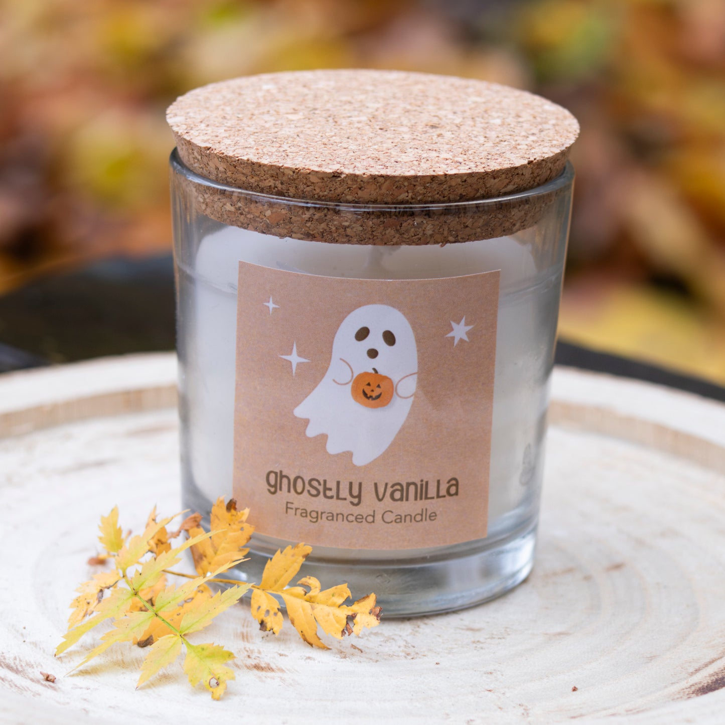 Eine weiße Duftkerze in einem klaren Glasbehälter mit einem naturbelassenen Korkdeckel. Das Etikett zeigt ein niedliches Geisterdesign, das einen kleinen Kürbis hält, sowie den Schriftzug ‘Ghostly Vanilla Fragranced Candle’. Die Kerze steht auf einer Holzscheibe, die mit herbstlichen Blättern dekoriert ist, und schafft eine gemütliche und saisonale Atmosphäre.