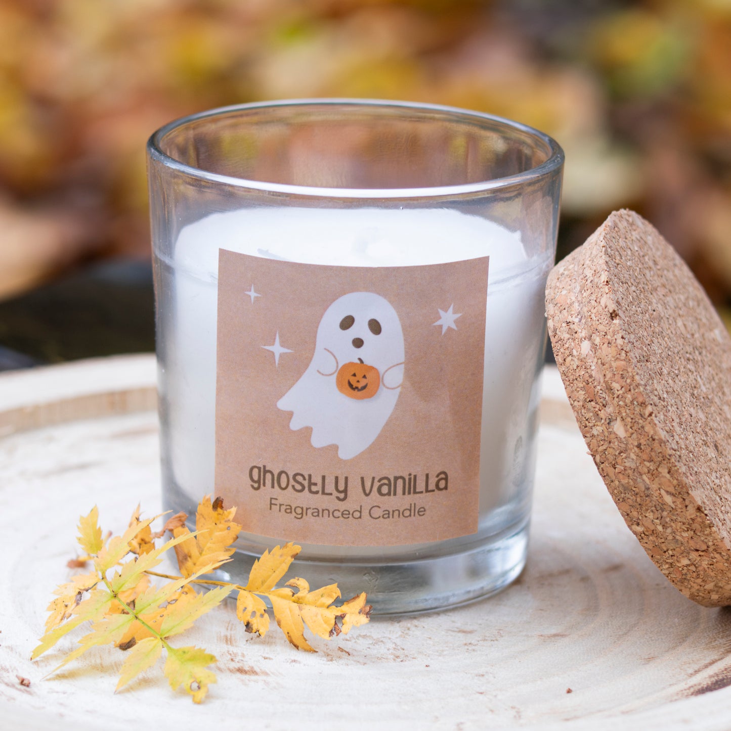 Eine zarte Duftkerze in einem klaren Glasbehälter, dekoriert mit einem Etikett, das ein niedliches Geisterdesign zeigt. Das Motiv ‘Ghostly Vanilla’ und der beige Hintergrund verleihen der Kerze eine herbstliche, verspielte Note. Die Kerze ist mit einem natürlichen Korkdeckel abgedeckt und auf einer Holzscheibe arrangiert, umgeben von herbstlichen Blättern, die eine warme und gemütliche Atmosphäre erzeugen.