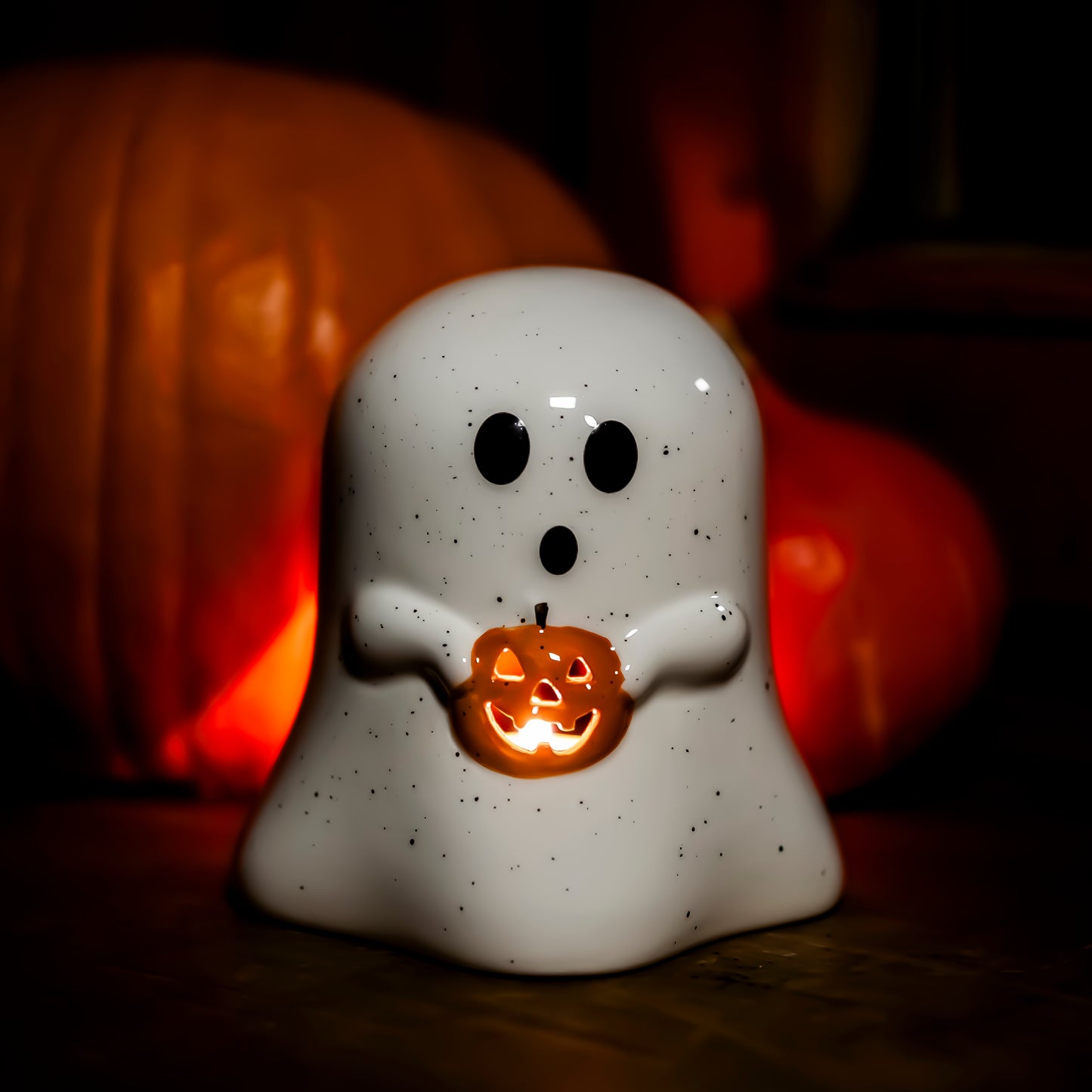 Eine weiße, gesprenkelte Geisterfigur mit großen schwarzen Augen, die einen leuchtenden Kürbis mit geschnitztem Gesicht hält. Im Hintergrund sind unscharfe orangefarbene Kürbisse zu sehen, die eine warme, herbstliche Atmosphäre schaffen.