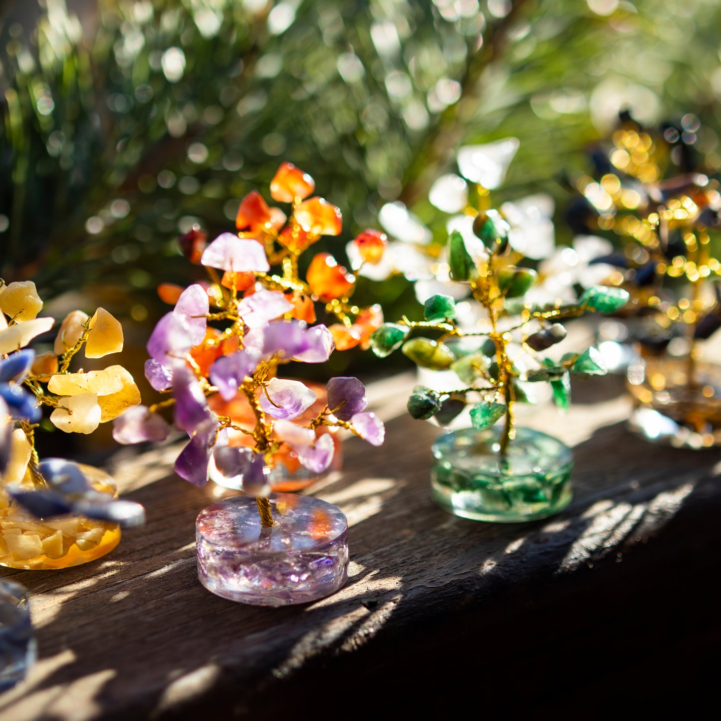 Verschiedene dekorative Glücksbäumchen aus bunten Halbedelsteinen und goldfarbenem Draht auf einer sonnigen Holzoberfläche, umgeben von einem natürlichen, unscharfen Hintergrund.