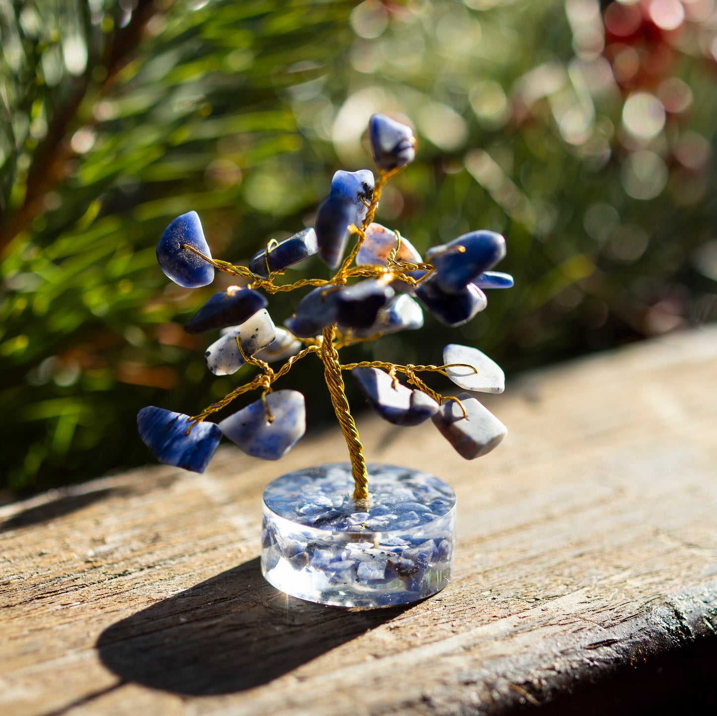 Dekoratives Bäumchen aus Lapislazuli-Steinen mit goldfarbenem Draht auf einer sonnigen Holzoberfläche, umgeben von unscharfem, grünem Hintergrund.