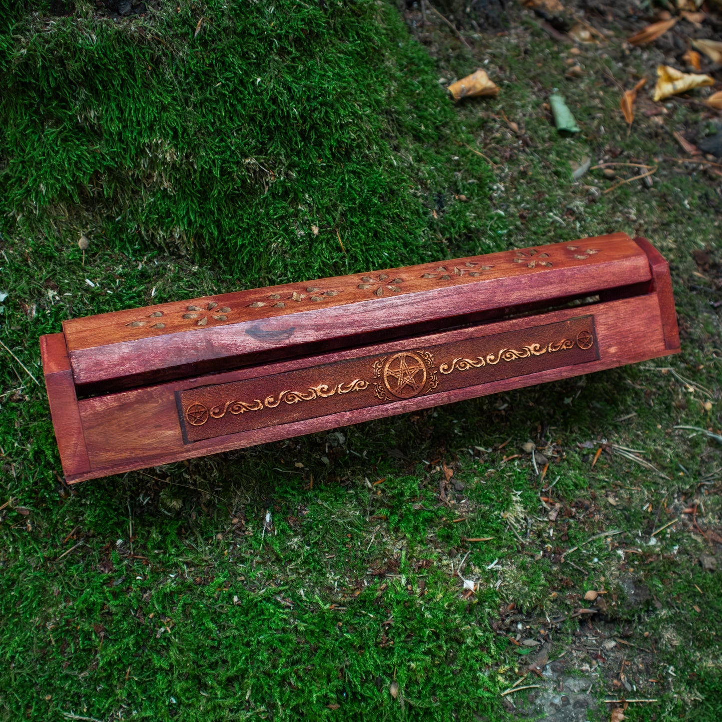 Ein Räucherstäbchenhalter aus Holz mit kunstvollen Verzierungen auf einem moosbedeckten Untergrund platziert, umgeben von natürlicher Vegetation.