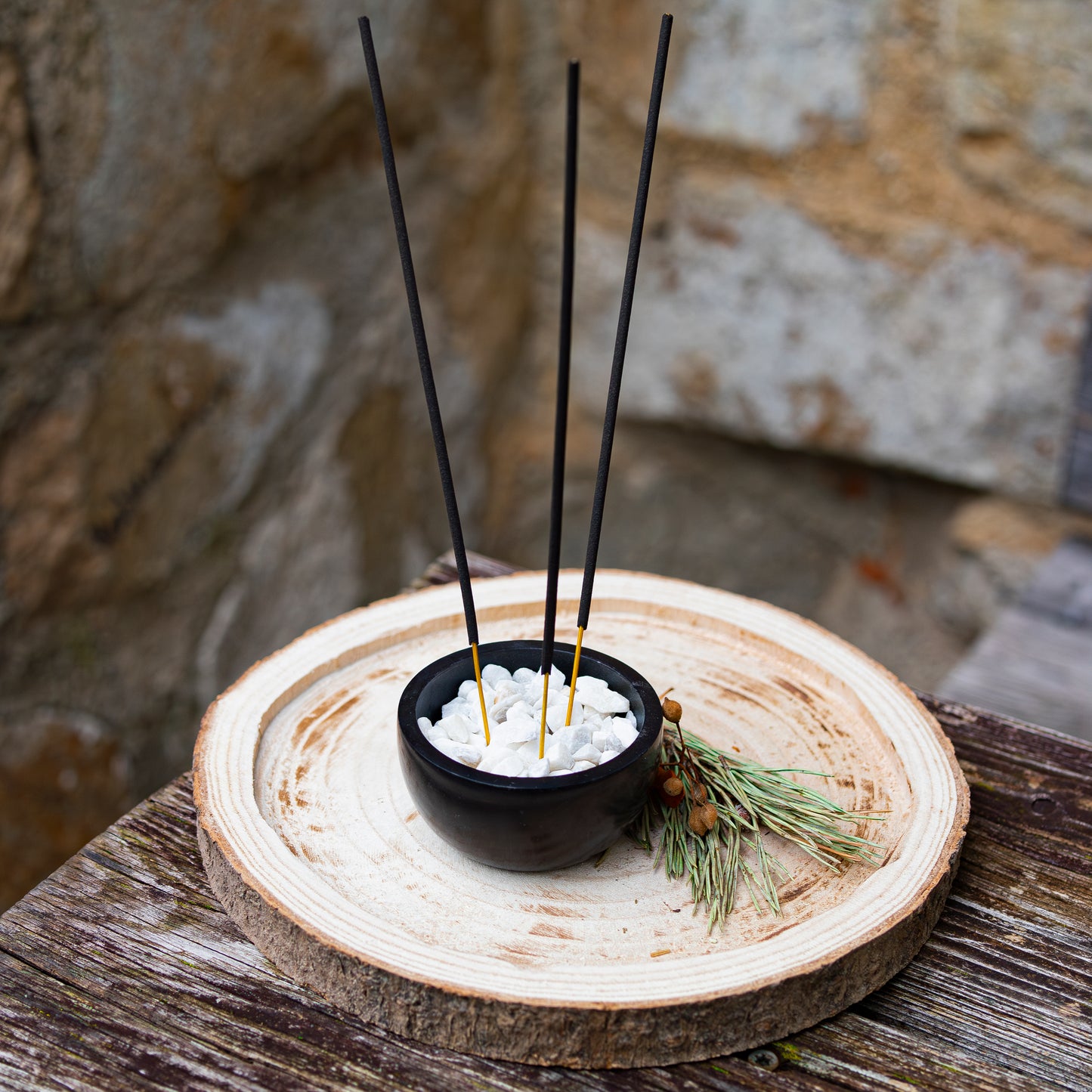 Schwarze Schale mit weißen Kieseln, in der drei Räucherstäbchen stehen, dekorativ auf einer Holzscheibe platziert und mit Tannenzweigen und Beeren verziert, vor einer rustikalen Steinwand.
