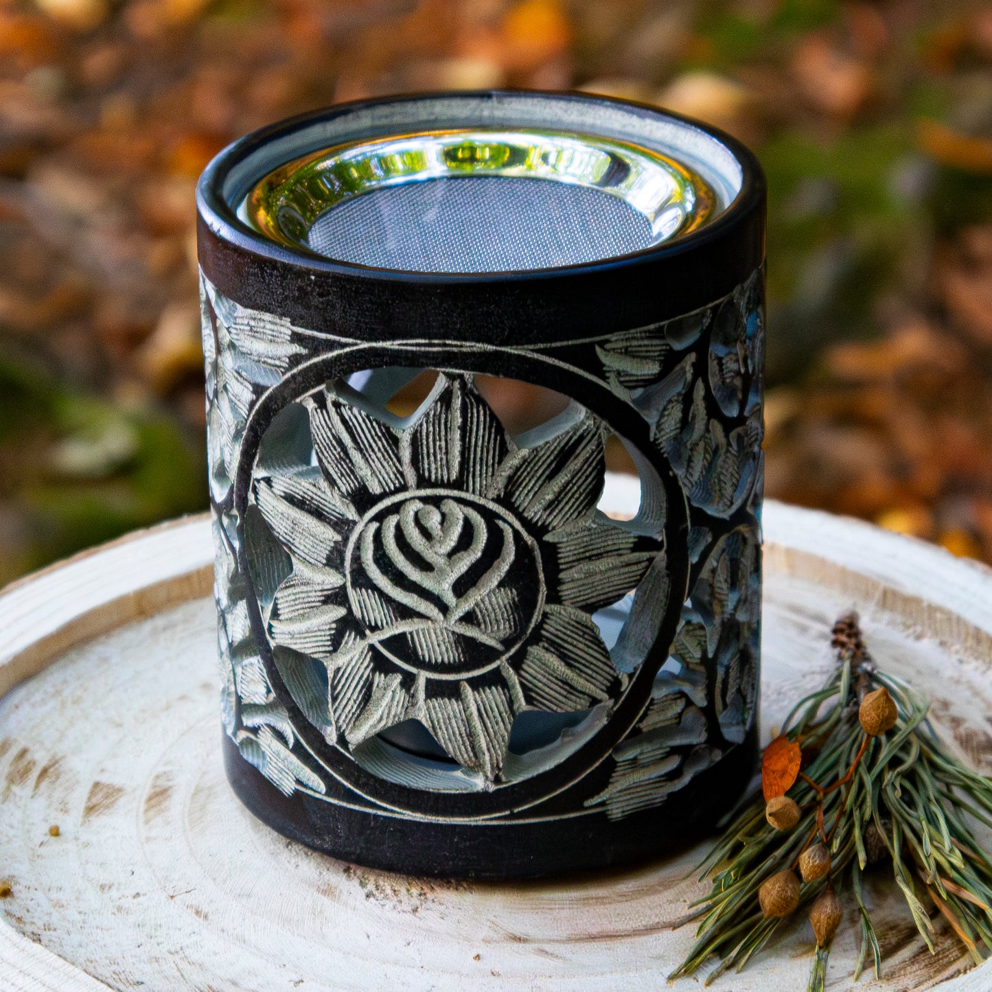Schwarzer Specksteinhalter mit filigranem Blumenmotiv, auf einer Holzscheibe platziert, mit dekorativem Zweig im Hintergrund.