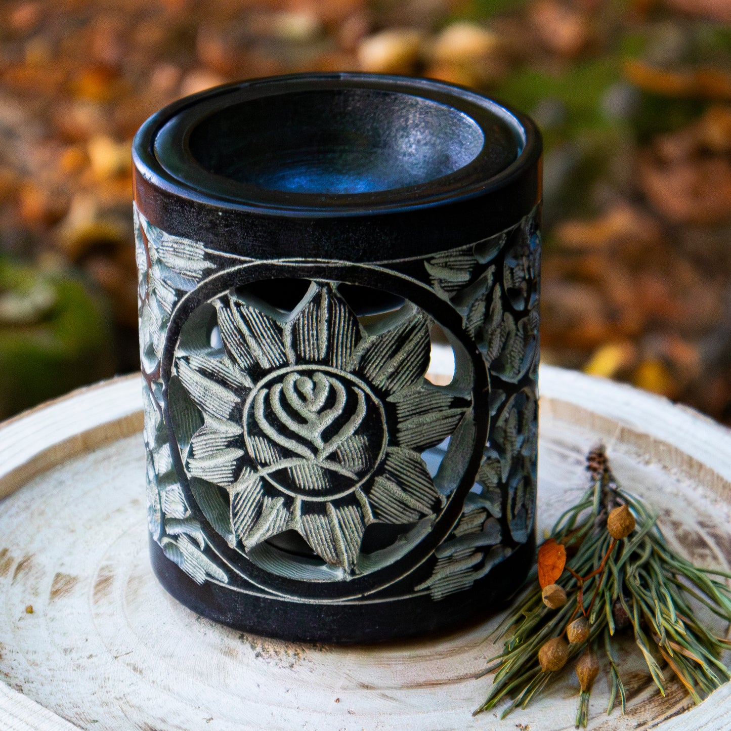 Schwarzer Specksteinhalter mit filigranem Blumenmotiv, auf einer Holzscheibe platziert, mit dekorativem Zweig im Hintergrund.