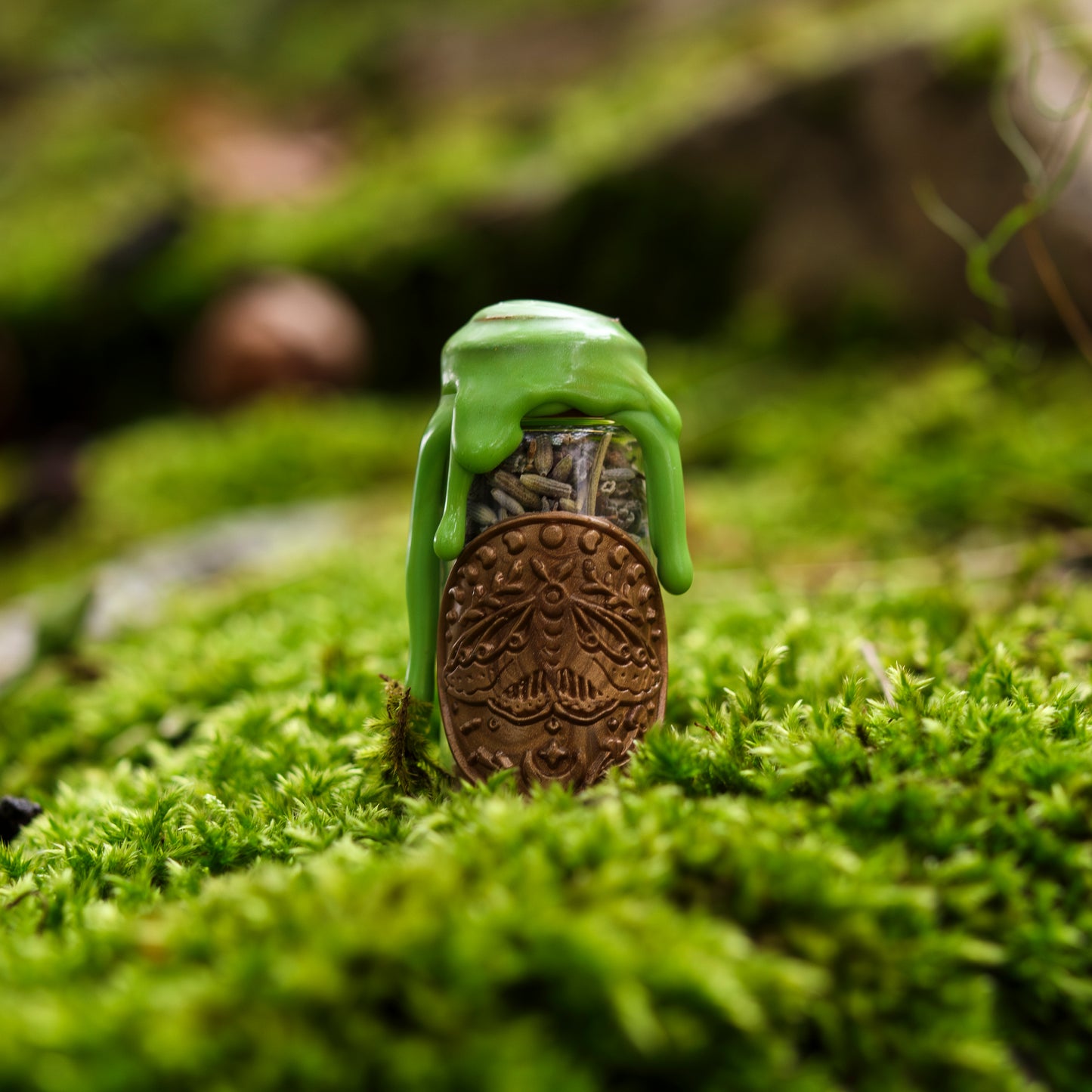 Kleine Spelljar ‚Glück‘ mit grüner Wachsversiegelung und einem dekorativen Anhänger mit geprägtem Motiv, auf moosbedecktem Untergrund präsentiert.