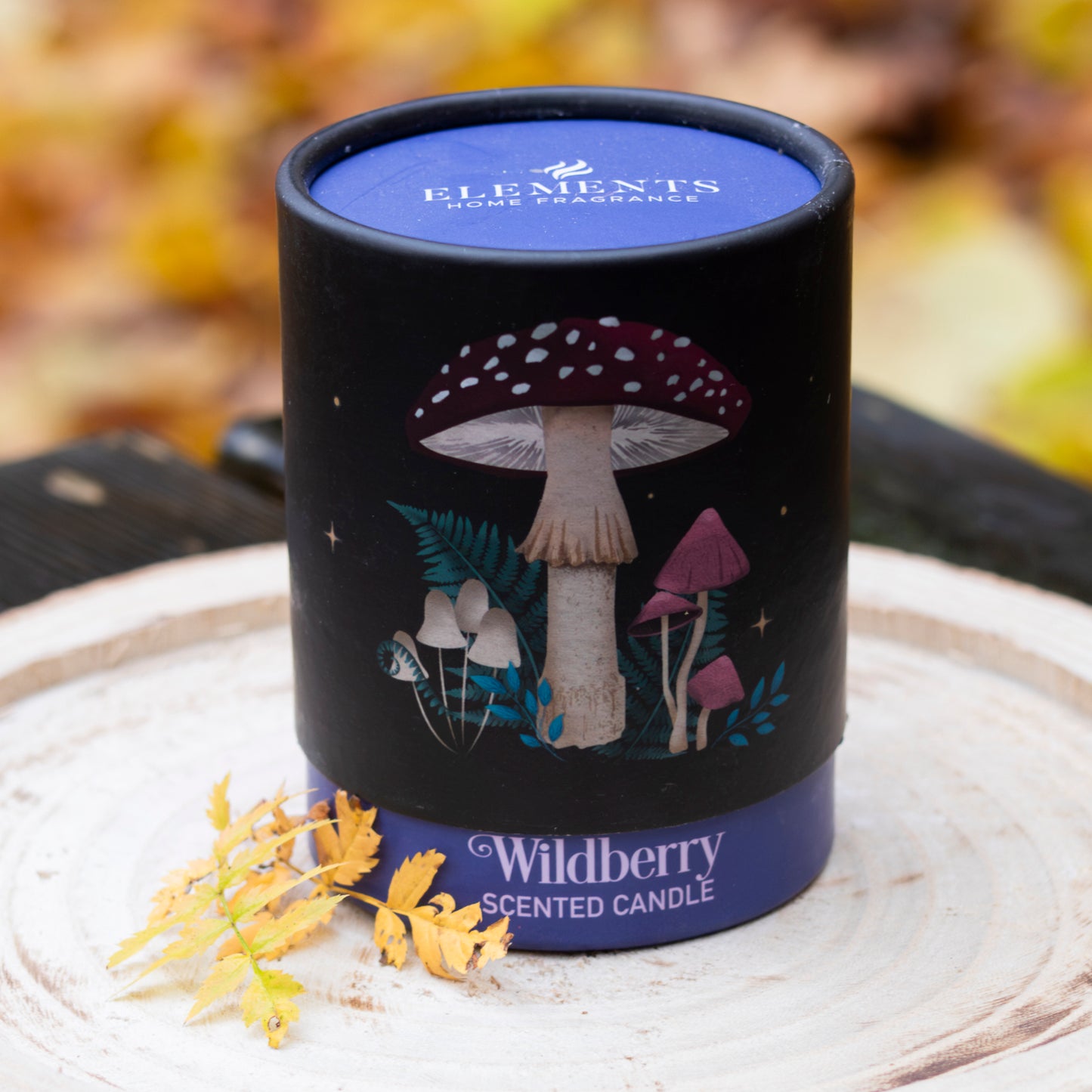 Duftkerze ‚Wildberry‘ in einer dekorativen Verpackung mit Pilzmotiv, auf einer Holzscheibe platziert und mit Herbstblättern verziert.