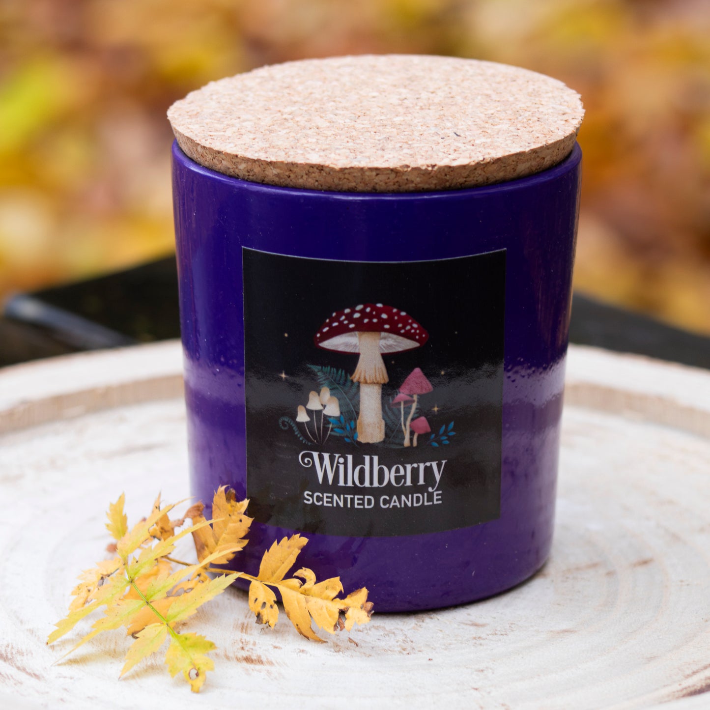 Lila Duftkerze mit der Aufschrift ‘Wildberry Scented Candle’, verziert mit einem Pilzdesign und einem Korkdeckel, dekoriert mit Herbstblättern auf einem rustikalen Holztablett.