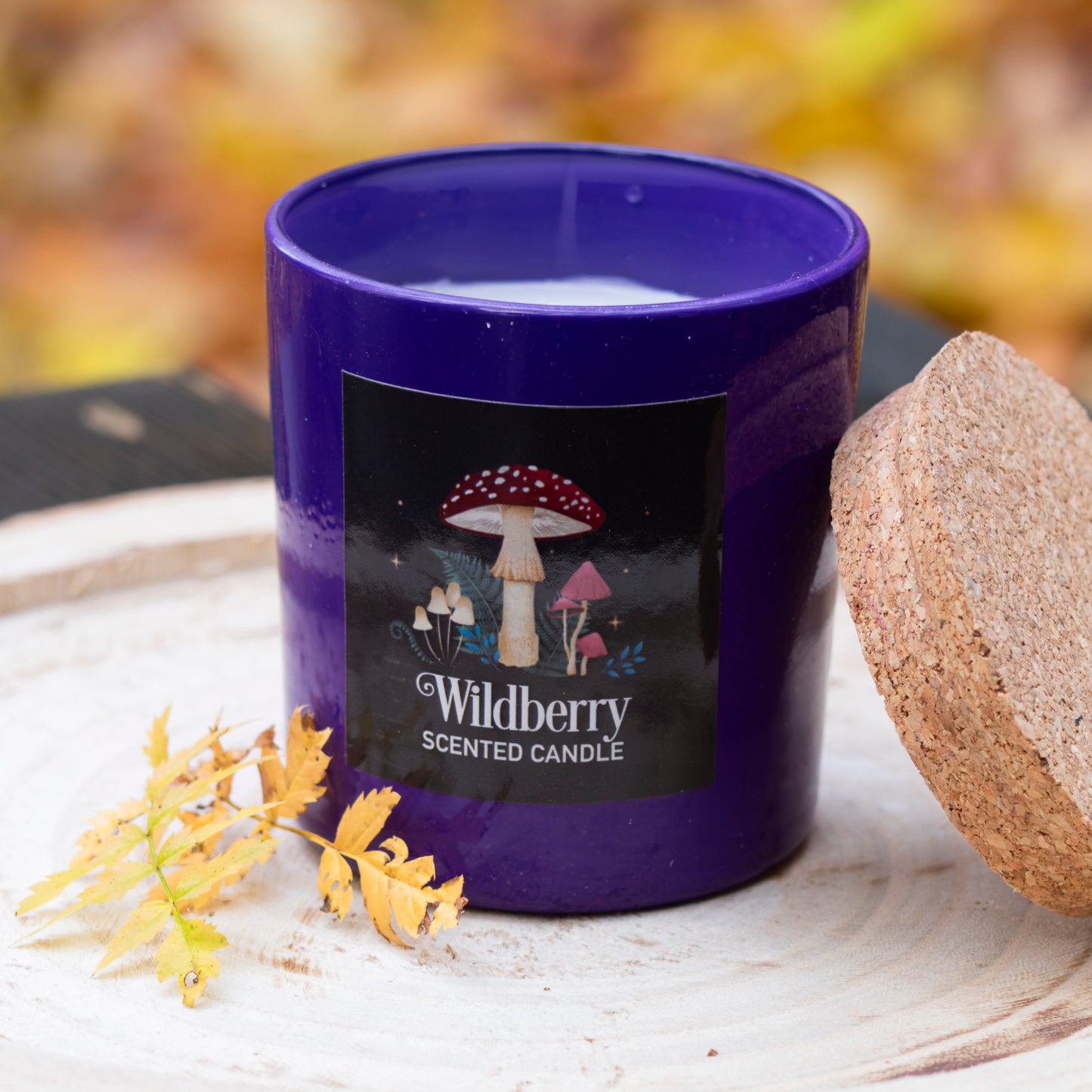 Eine tiefviolette Duftkerze in einem eleganten Glasbehälter mit einem naturbelassenen Korkdeckel. Das Etikett zeigt ein stimmungsvolles Design mit Waldpilzen und Pflanzen sowie dem Schriftzug ‘Wildberry Scented Candle’. Die Kerze steht auf einer rustikalen Holzscheibe, dekoriert mit herbstlichen Blättern, und vermittelt eine warme, naturverbundene Atmosphäre.