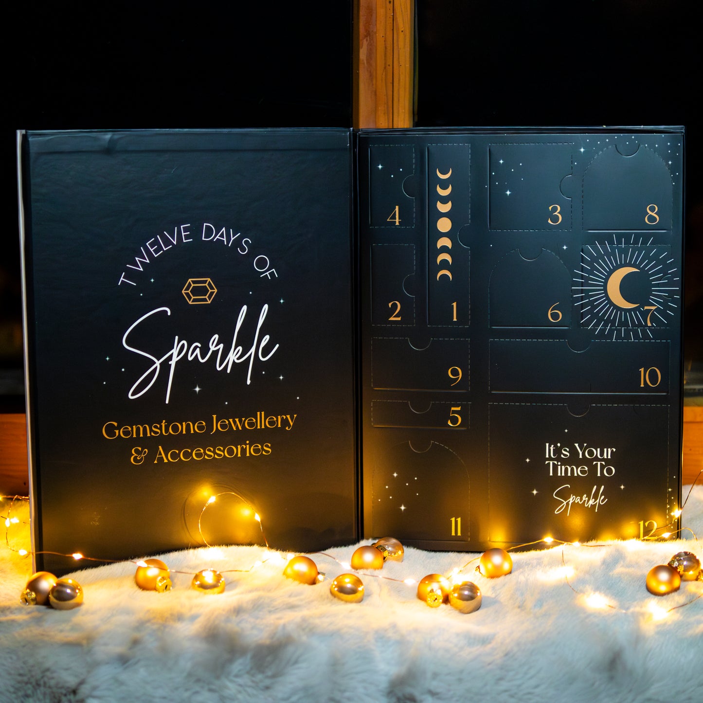 Ein edler Adventskalender in Schwarz mit goldener Aufschrift ‘Twelve Days of Sparkle – Gemstone Jewellery & Accessories’. Der Kalender zeigt nummerierte Türchen mit Symbolen von Monden und Sternen. Im Vordergrund liegen funkelnde Lichter und goldene Kugeln auf einer weichen weißen Oberfläche.