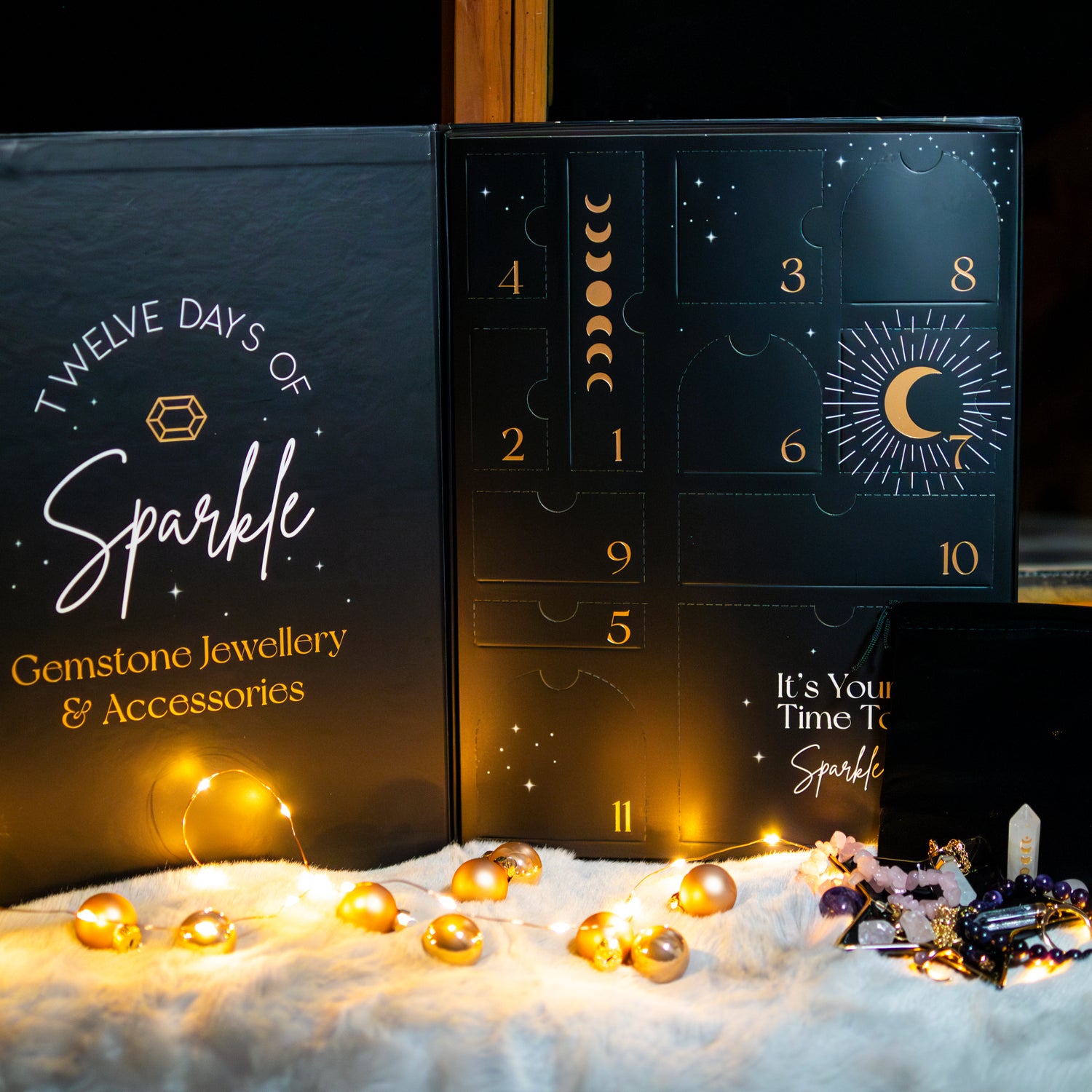 Ein edler Adventskalender in Schwarz mit goldener Aufschrift ‘Twelve Days of Sparkle – Gemstone Jewellery & Accessories’. Der Kalender zeigt nummerierte Türchen mit Symbolen von Monden und Sternen. Im Vordergrund liegen funkelnde Lichter, goldene Kugeln und dekorative Edelsteine auf einer weichen weißen Oberfläche.