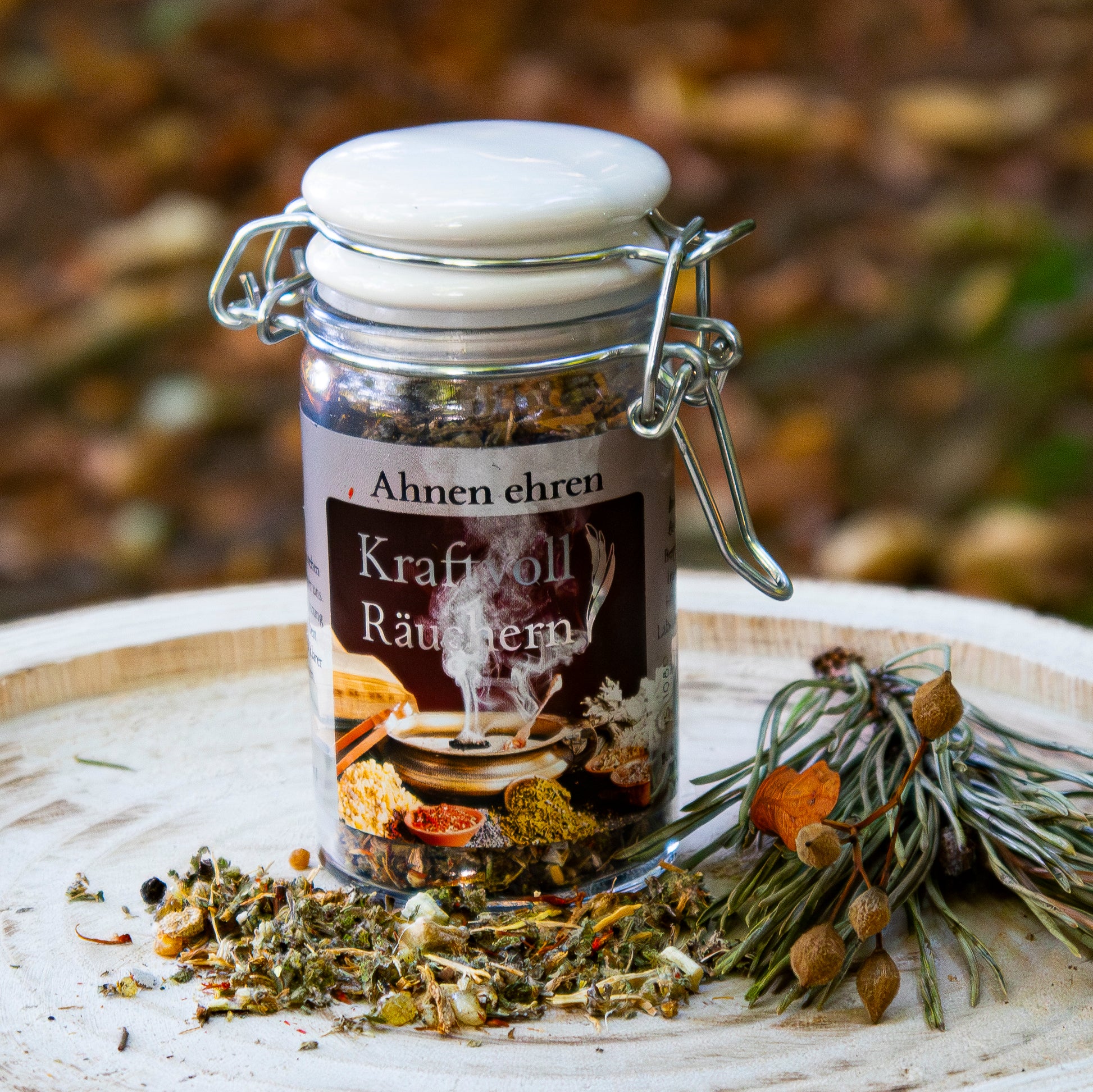 Glasgefäß mit der Aufschrift ‘Ahnen ehren – Kraftvoll Räuchern’, gefüllt mit einer Kräutermischung, arrangiert auf einer Holzscheibe mit losem Räucherwerk und getrockneten Zweigen vor herbstlicher Kulisse.