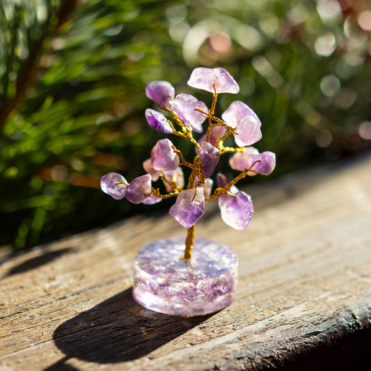 Dekoratives Bäumchen aus Amethyst-Steinen mit goldfarbenem Draht auf einer sonnigen Holzoberfläche, umgeben von unscharfem, grünem Hintergrund.