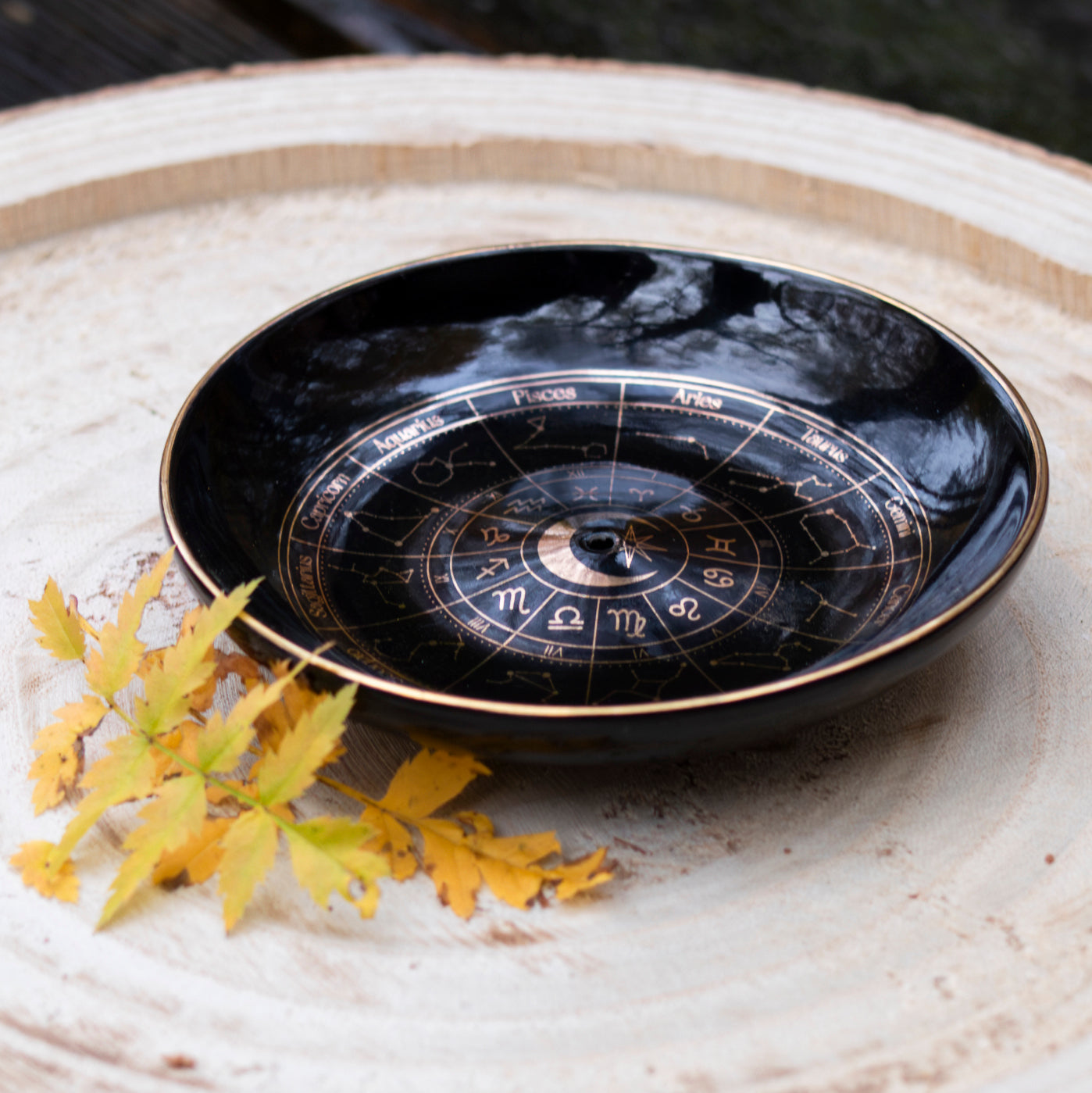 Astrologie-Räucherstäbchenschale mit goldenen Tierkreiszeichen auf schwarzem Hintergrund, dekorativ auf einer Holzscheibe platziert und mit Herbstblättern verziert.