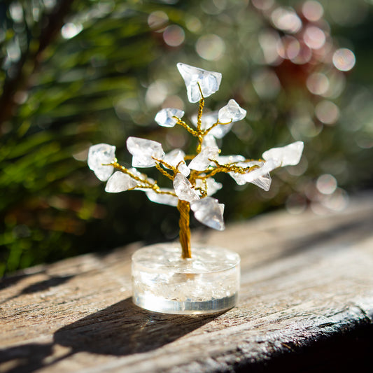 Dekoratives Bäumchen aus Bergkristall-Steinen mit goldfarbenem Draht auf einer sonnigen Holzoberfläche, umgeben von unscharfem, grünem Hintergrund.