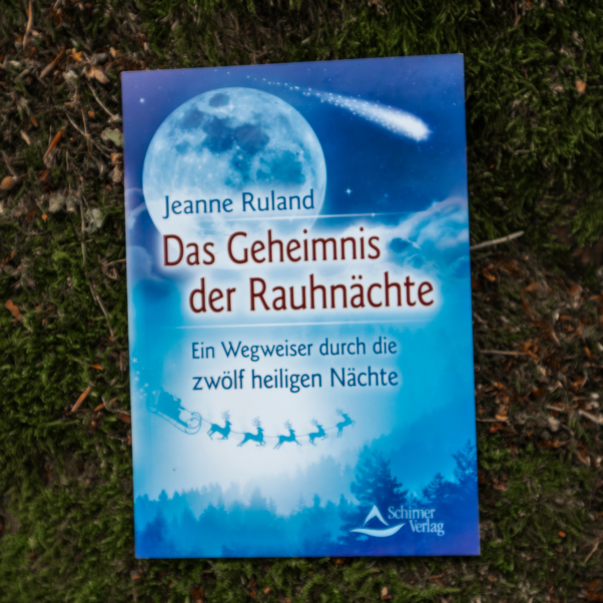 Das Buch ‘Das Geheimnis der Rauhnächte’ von Jeanne Ruland, mit einem blauen Cover, das einen leuchtenden Vollmond und eine winterliche Szenerie zeigt, auf einem moosbedeckten Waldboden liegend.