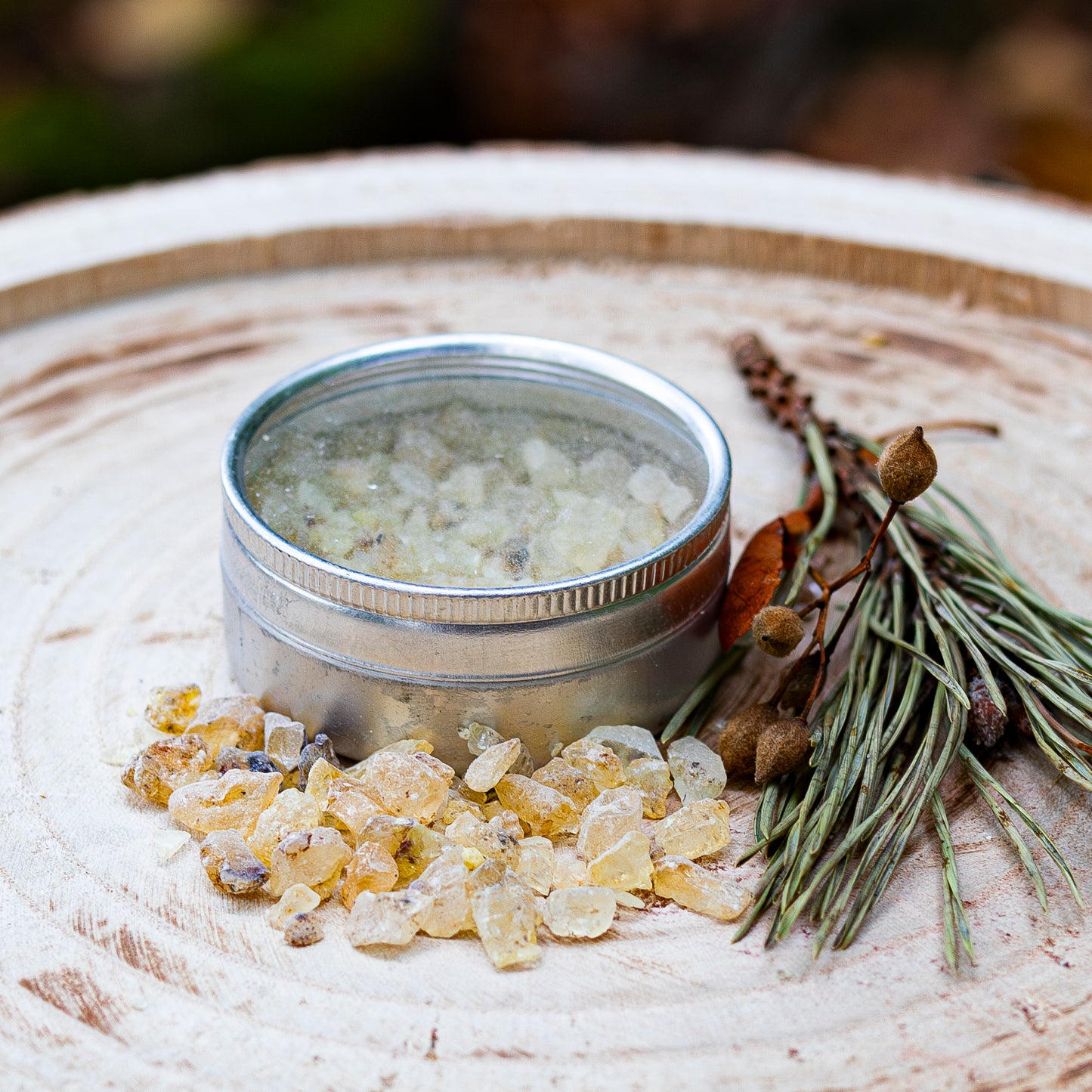 Silberne Dose mit geöffnetem Deckel, gefüllt mit Copal-Harz, arrangiert auf einer Holzscheibe mit verstreuten Harzstücken und einem dekorativen Bündel aus Tannenzweigen und getrockneten Pflanzen.