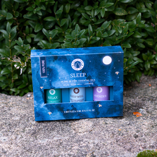 Ein ätherisches Öl-Set namens ‘Sleep’ in einer blauen, nachthimmel-inspierierten Verpackung, präsentiert auf einer Steinfläche vor grüner Hecke.