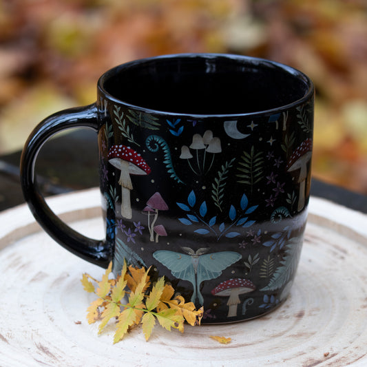 Eine schwarze Tasse mit Waldmotiv, verziert mit Illustrationen von Pilzen, Blättern, einem Nachtfalter und einem Mond. Die Tasse steht auf einer Holzscheibe, dekoriert mit gelben Herbstblättern. Der Hintergrund zeigt unscharfes herbstliches Laub.