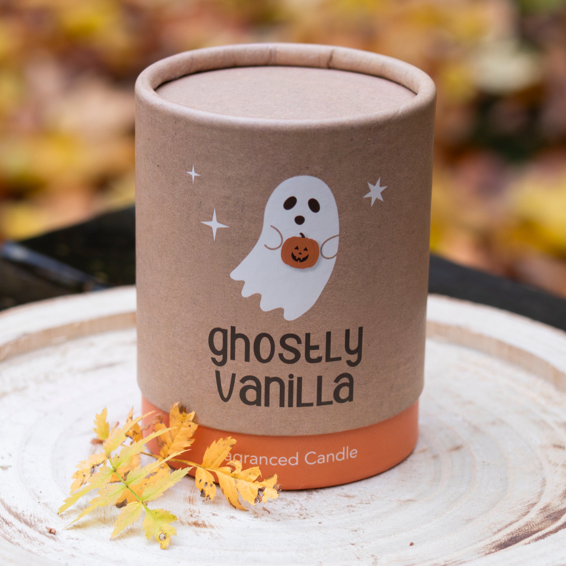 Eine dekorative Kerze mit der Aufschrift ‘Ghostly Vanilla’, verpackt in einem zylindrischen Karton mit einem niedlichen Geist, der einen Kürbis hält. Der Hintergrund zeigt herbstliche Blätter, passend zur saisonalen Stimmung.