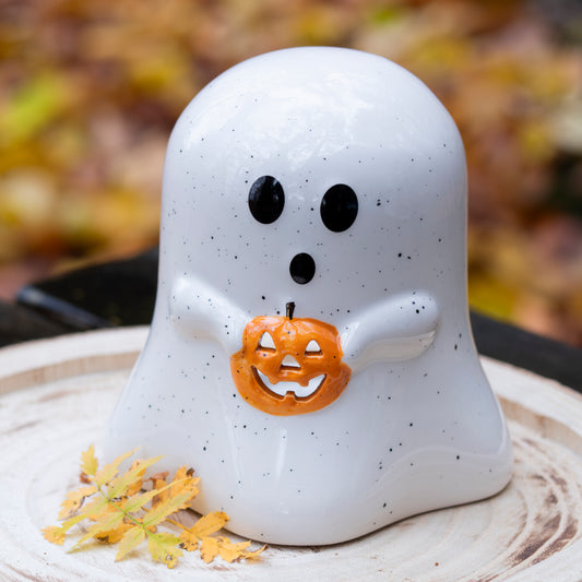 Keramikgeist mit schwarzen Augen und Mund, der einen kleinen Kürbis mit Gesicht hält, auf einem rustikalen Holztablett mit Herbstblättern.