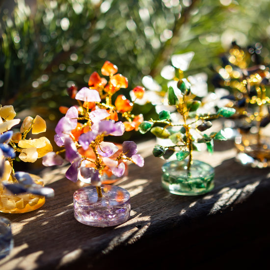 Verschiedene dekorative Glücksbäumchen aus bunten Halbedelsteinen und goldfarbenem Draht auf einer sonnigen Holzoberfläche, umgeben von einem natürlichen, unscharfen Hintergrund.