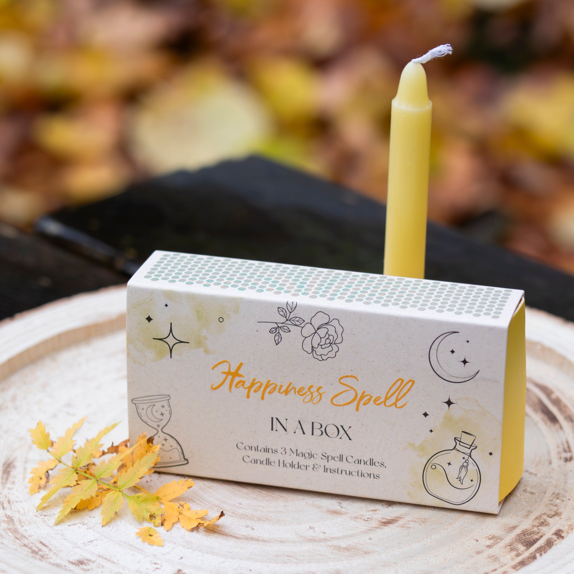 Eine Box mit der Aufschrift ‘Happiness Spell in a Box’, verziert mit Illustrationen von Blumen, Sternen und einem Mond. Auf der Box steht eine gelbe Kerze. Die Szene ist auf einer Holzscheibe arrangiert und mit gelben Herbstblättern dekoriert. Der Hintergrund zeigt unscharfes herbstliches Laub.