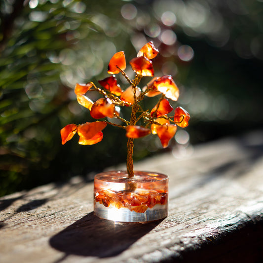 Dekoratives Bäumchen aus Karneol-Steinen mit goldfarbenem Draht auf einer sonnigen Holzoberfläche, umgeben von unscharfem, grünem Hintergrund.