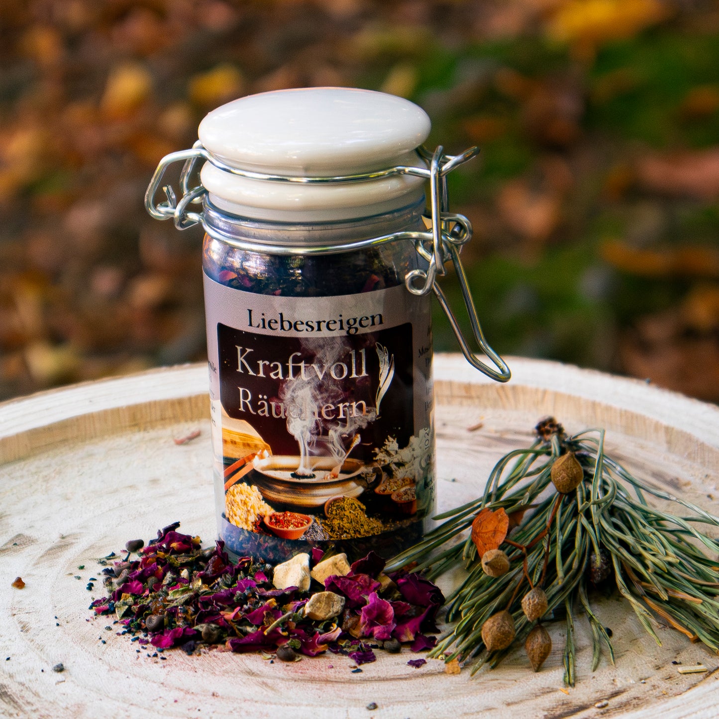 Glasbehälter mit der Aufschrift ‘Liebesreigen – Kraftvoll Räuchern’, gefüllt mit Räuchermischung, auf einer Holzscheibe arrangiert, dekoriert mit getrockneten Blüten und Kräutern vor herbstlichem Hintergrund.