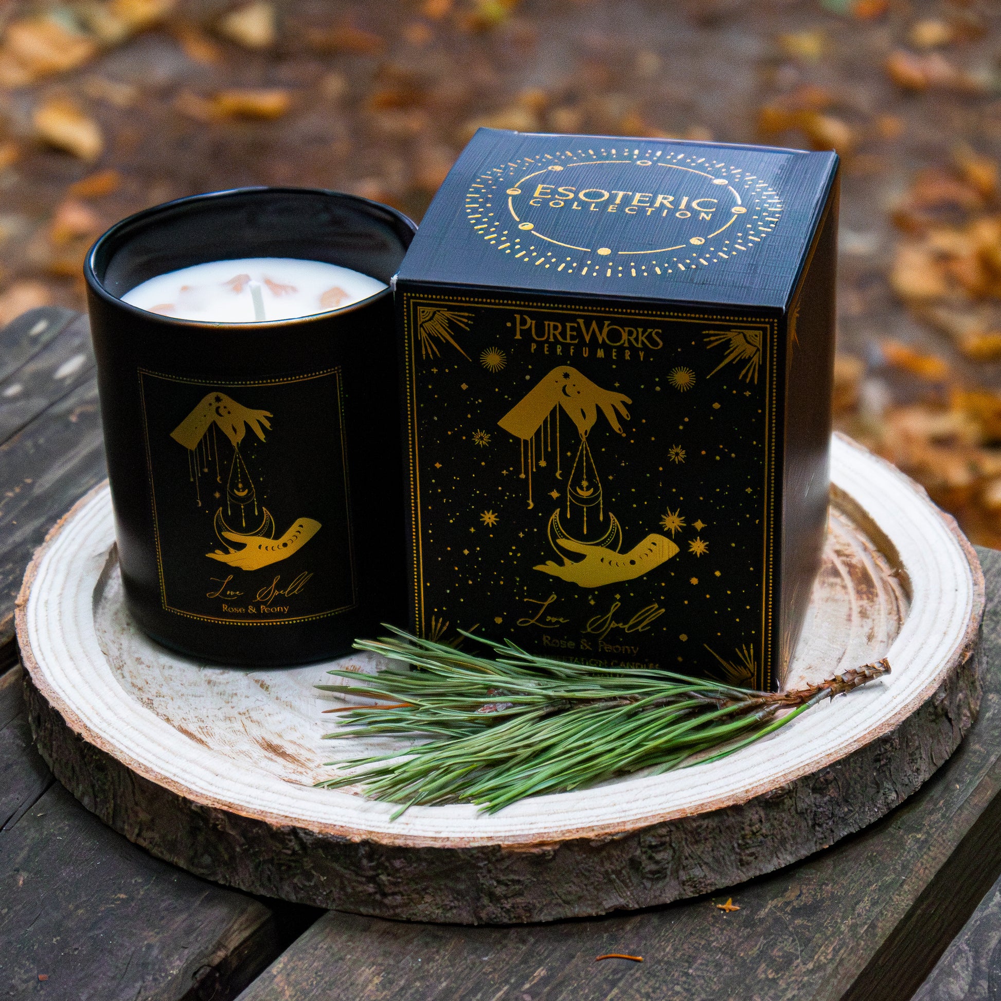 Duftkerze ‚Love Spell‘ in schwarzer Dose mit goldenen Verzierungen, neben passender Verpackung auf einer Holzscheibe arrangiert und mit Zweigen dekoriert, vor herbstlicher Kulisse.