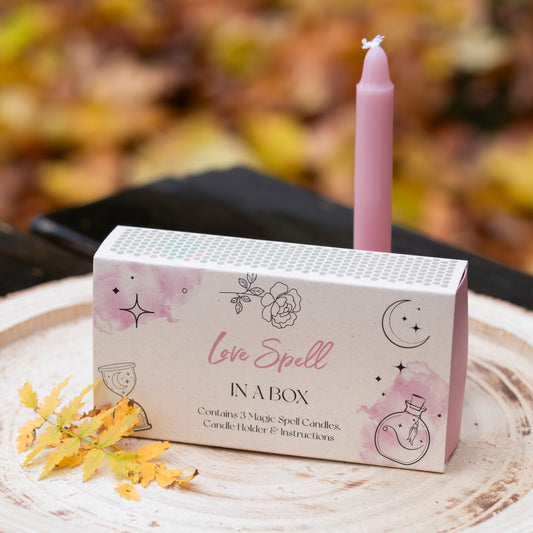 Eine Box mit der Aufschrift ‘Love Spell in a Box’, dekoriert mit Illustrationen von Blumen, Sternen und einem Mond. Auf der Box steht eine rosafarbene Kerze. Die Szene ist auf einer Holzscheibe arrangiert, dekoriert mit gelben Herbstblättern. Der Hintergrund zeigt unscharfes herbstliches Laub.