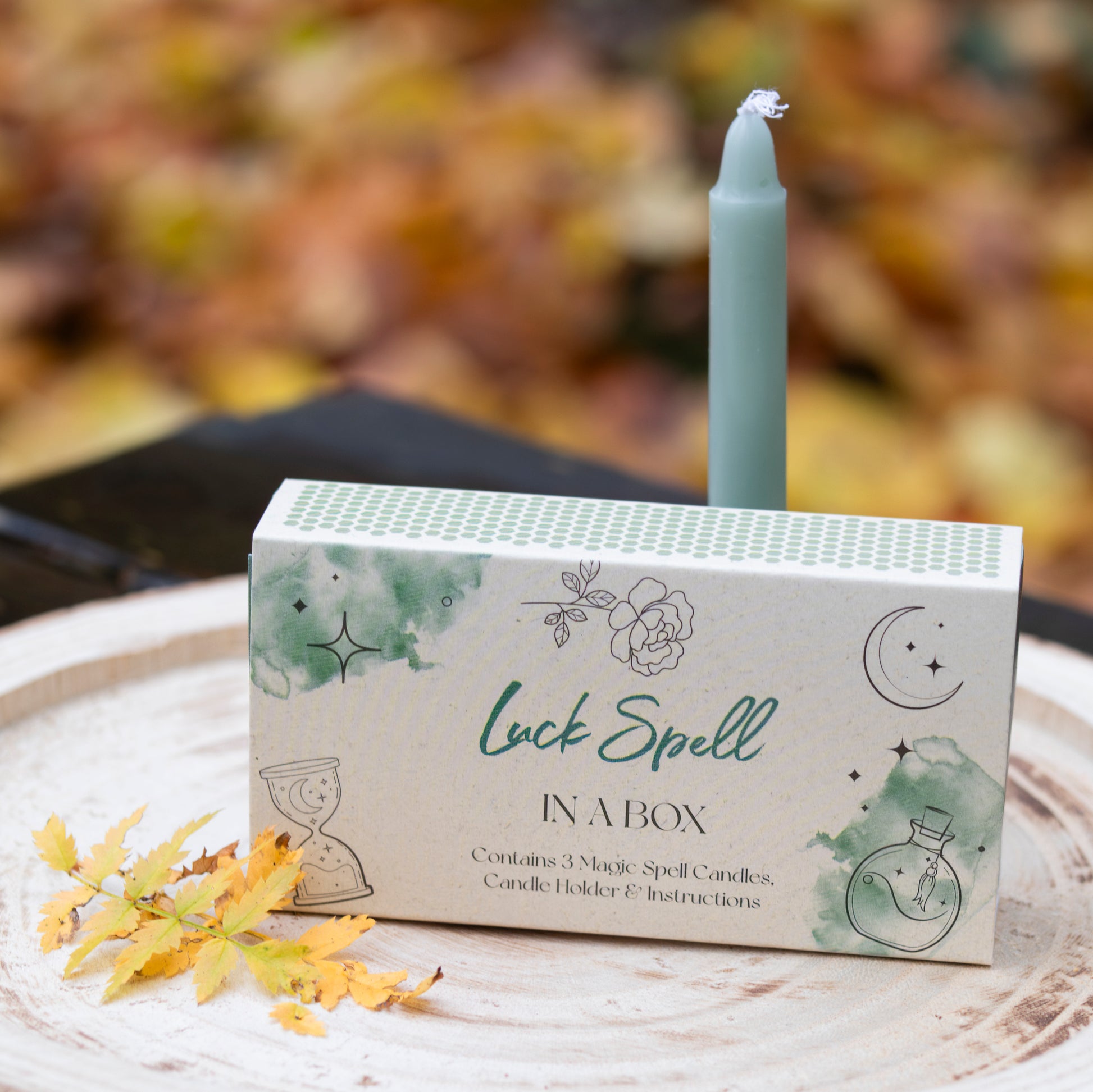 Eine Box mit der Aufschrift ‘Luck Spell in a Box’, dekoriert mit Illustrationen von Blumen, einem Mond und einer Sanduhr. Auf der Box steht eine grüne Kerze. Die Szene ist auf einer runden Holzscheibe arrangiert und mit gelben Herbstblättern dekoriert. Im Hintergrund sind unscharfe herbstliche Blätter zu sehen.