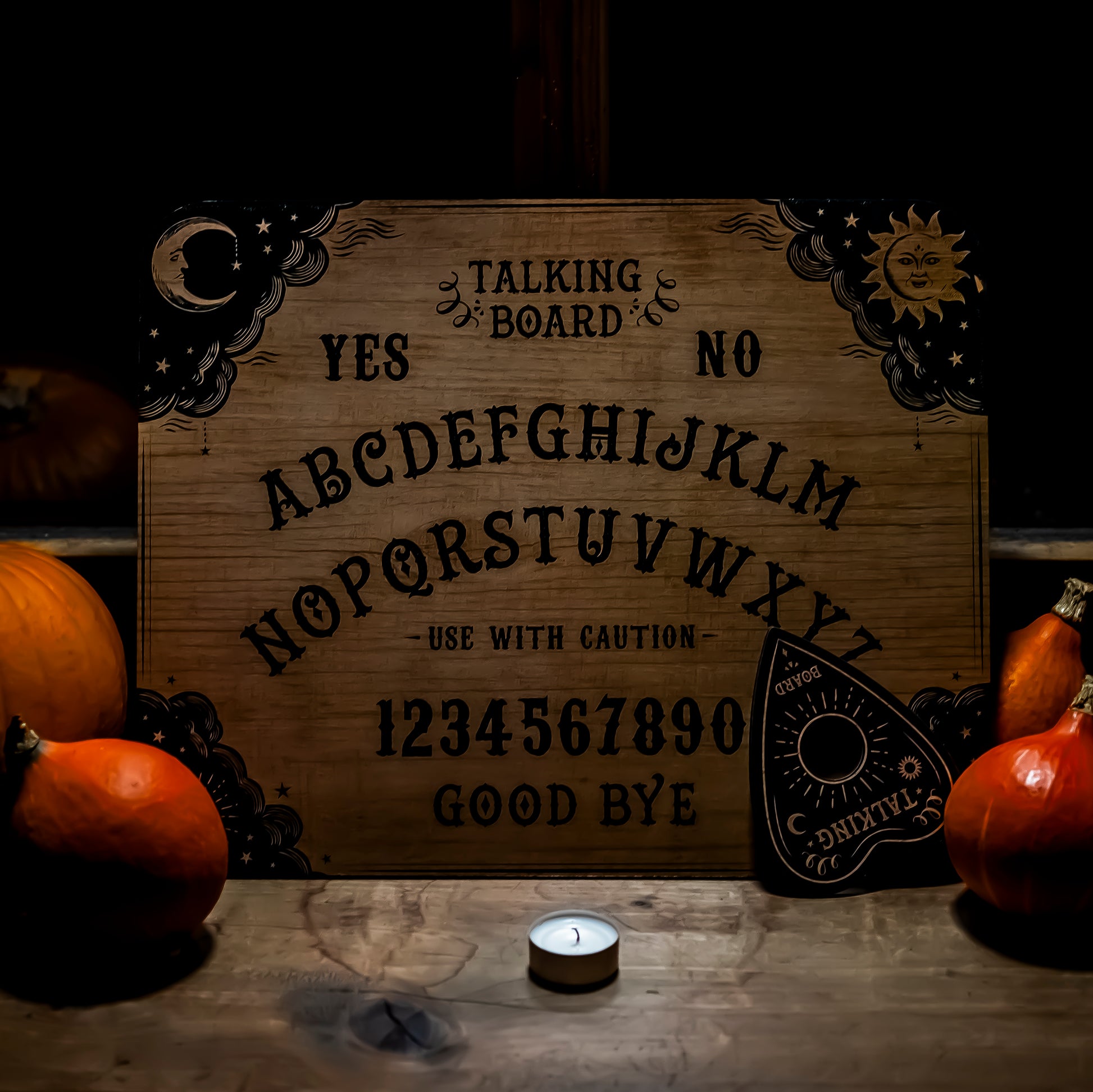 Ein klassisches Ouija-Brett aus Holz mit detaillierten Verzierungen von Sonne und Mond in den Ecken. Die Buchstaben des Alphabets sowie Zahlen und Wörter wie ‘Yes’, ‘No’ und ‘Good Bye’ sind auf das Brett graviert. Im Vordergrund ein kleines Teelicht, umgeben von dekorativen Kürbissen, die eine herbstliche Stimmung erzeugen.