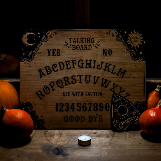 Ein klassisches Ouija-Brett aus Holz mit detaillierten Verzierungen von Sonne und Mond in den Ecken. Die Buchstaben des Alphabets sowie Zahlen und Wörter wie ‘Yes’, ‘No’ und ‘Good Bye’ sind auf das Brett graviert. Im Vordergrund ein kleines Teelicht, umgeben von dekorativen Kürbissen, die eine herbstliche Stimmung erzeugen.