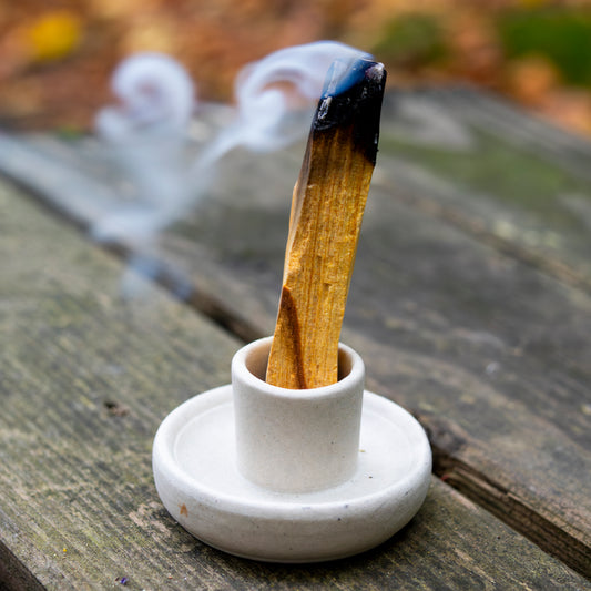 Brennendes Palo-Santo-Holzstück, aufrecht in einem schlichten weißen Halter arrangiert, auf einer Holzoberfläche vor unscharfem, herbstlichem Hintergrund.