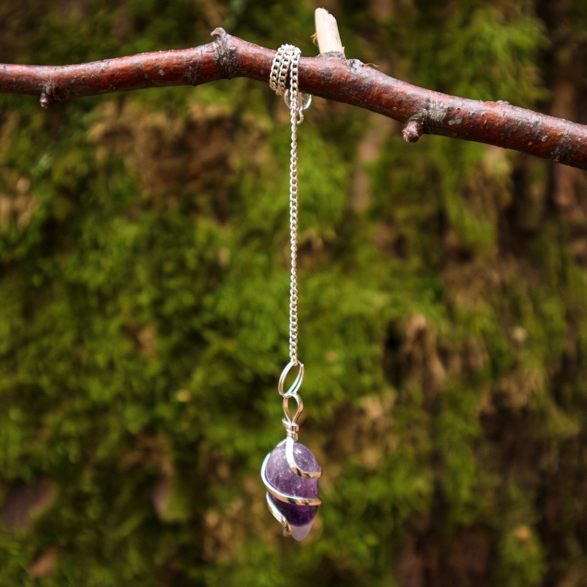 Pendel mit einem Amethyst-Stein, eingewickelt in Silberdraht, an einer silbernen Kette hängend, dekorativ an einem Ast vor einem moosbewachsenen Hintergrund präsentiert.