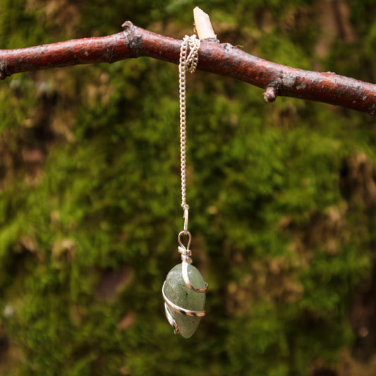 Pendel mit einem Aventurin-Stein, eingewickelt in Silberdraht, an einer silbernen Kette hängend, dekorativ an einem Ast vor einem moosbewachsenen Hintergrund präsentiert.