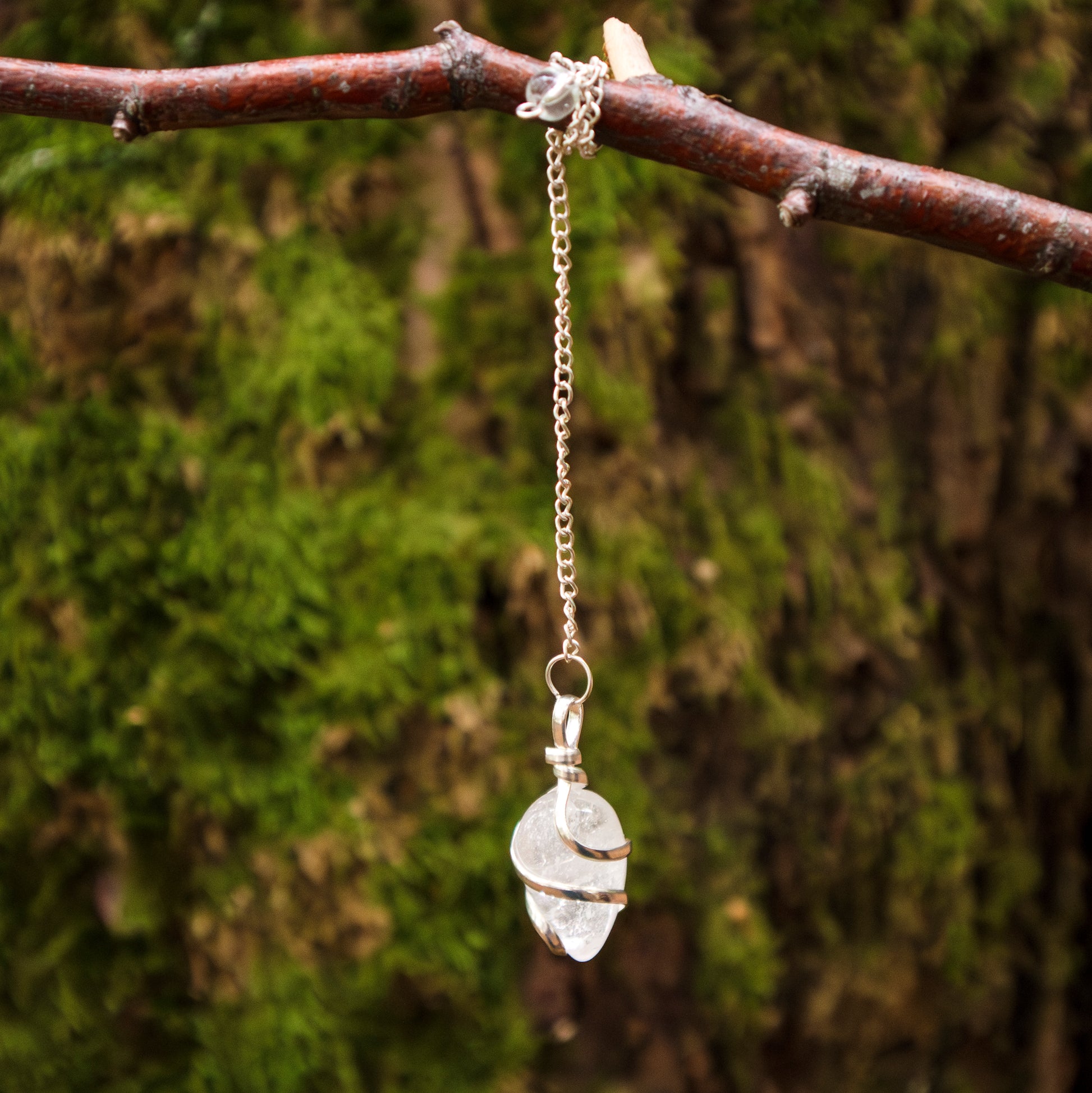 Pendel mit einem Bergkristall-Stein, eingewickelt in Silberdraht, an einer silbernen Kette hängend, dekorativ an einem Ast vor einem moosbewachsenen Hintergrund präsentiert.