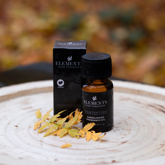 Eine kleine braune Flasche mit der Aufschrift ‘Elements Home Fragrance - Protection Sandalwood Fragrance Oil’, begleitet von einer passenden schwarzen Verpackung. Die Flasche steht auf einer runden Holzscheibe, dekoriert mit gelben Herbstblättern. Der Hintergrund zeigt unscharfes herbstliches Laub.