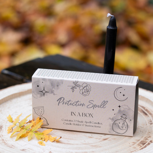 Eine Box mit der Aufschrift ‘Protection Spell in a Box’, verziert mit Illustrationen von Blumen, Sternen und einem Mond. Auf der Box steht eine schwarze Kerze. Die Szene ist auf einer Holzscheibe arrangiert und mit gelben Herbstblättern dekoriert. Der Hintergrund zeigt unscharfes herbstliches Laub.