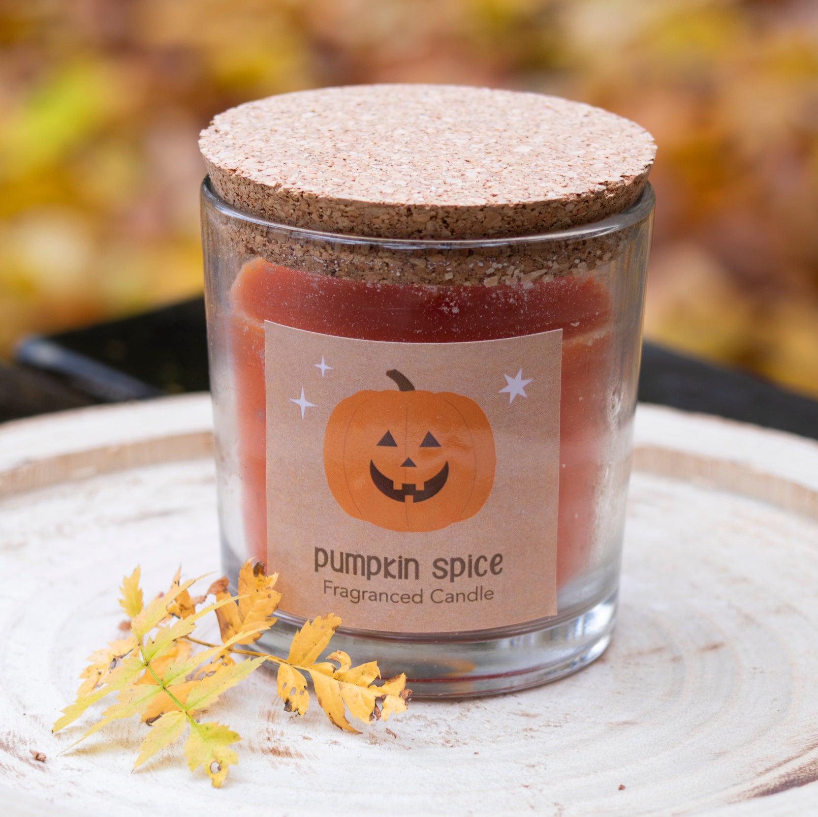 Eine duftende Kerze im Glas mit der Aufschrift ‘Pumpkin Spice’ und einem fröhlichen Kürbis-Motiv auf dem Etikett. Die Kerze hat eine warme orangefarbene Farbe und wird von einem passenden korkähnlichen Deckel verschlossen. Der Hintergrund zeigt herbstliche Blätter, die die gemütliche und saisonale Atmosphäre unterstreichen.