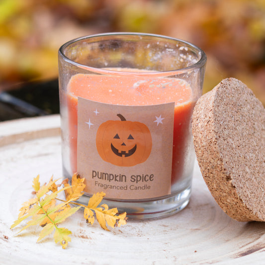 Eine duftende Kerze im Glas mit der Aufschrift ‘Pumpkin Spice’ und einem fröhlich grinsenden Kürbis-Motiv auf dem Etikett. Die Kerze hat eine warme orangefarbene Farbe, passend zum Herbstthema, und ist mit einem korkähnlichen Deckel ausgestattet, der seitlich liegt. Der Hintergrund zeigt herbstliche Blätter, die die saisonale Stimmung unterstreichen.