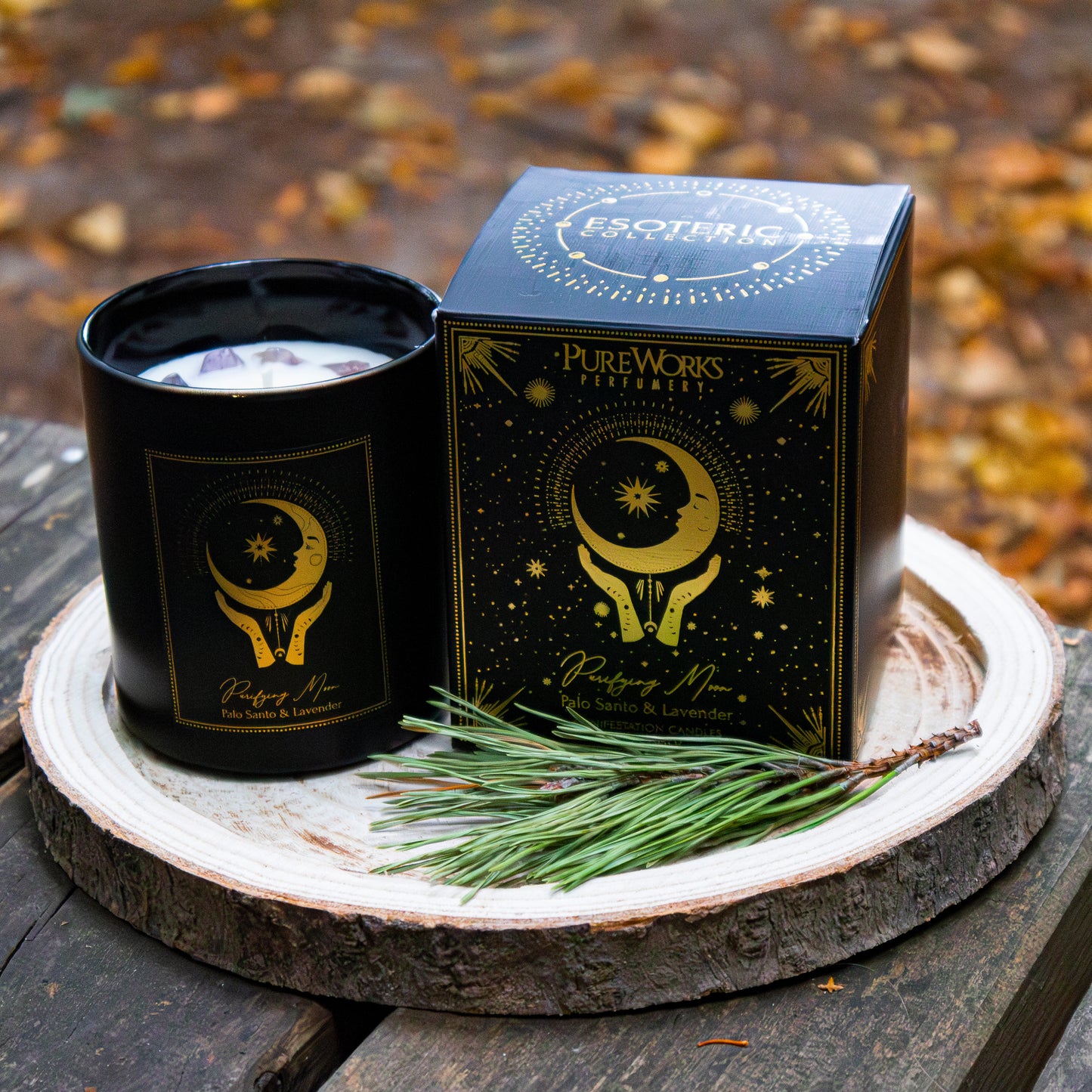 Duftkerze ‚Purifying Moon‘ in schwarzer Dose mit goldenen Verzierungen, neben passender Verpackung auf einer Holzscheibe arrangiert und mit Zweigen dekoriert, vor herbstlicher Kulisse.