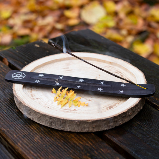 Ein schwarzes Räucherstäbchenhalter aus Holz mit Pentagramm- und Sternensymbolen, platziert auf einer runden Holzscheibe. Darauf brennt ein Räucherstäbchen, dessen Rauch sichtbar aufsteigt. Im Hintergrund herbstliche Blätter auf einer Holzbank.