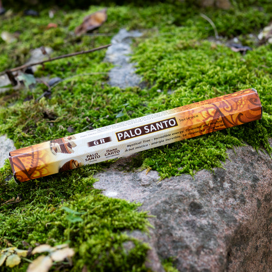 Verpackung von Palo Santo-Räucherstäbchen in warmen Braun- und Orangetönen, dekorativ auf einem moosbedeckten Stein in einer natürlichen Umgebung arrangiert.