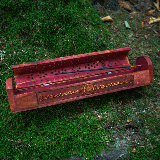 Ein Räucherstäbchenhalter aus Holz mit kunstvollen Verzierungen, geöffnet auf einem moosbedeckten Untergrund platziert, umgeben von natürlicher Vegetation.