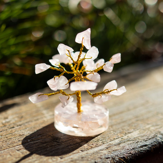 Dekoratives Bäumchen aus Rosenquarz-Steinen mit goldfarbenem Draht auf einer sonnigen Holzoberfläche, umgeben von unscharfem, grünem Hintergrund.
