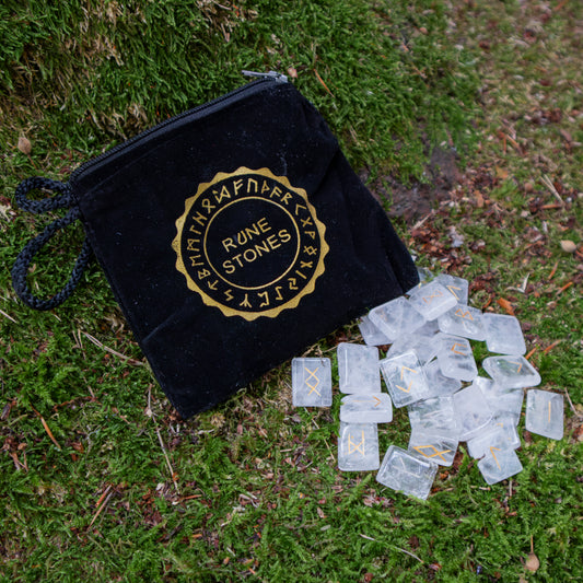 Ein schwarzer Stoffbeutel mit goldener Aufschrift ‘Rune Stones’, daneben transparente Runensteine mit goldenen Symbolen, liegend auf einer grünen Moosfläche.