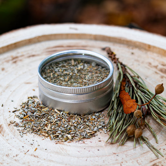 Runde Metall-Dose mit getrocknetem Salbei, daneben verstreute Salbeiblätter und ein dekoratives Arrangement aus Tannenzweigen und Beeren, auf einer Holzscheibe präsentiert.