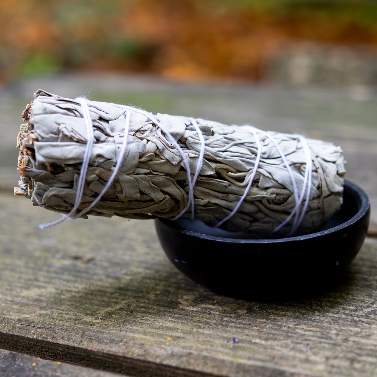 Weißer Salbei-Bündel, mit Schnur umwickelt, dekorativ in einer schwarzen Schale auf einer Holzoberfläche präsentiert, vor unscharfem herbstlichem Hintergrund.